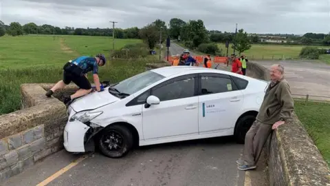 A white car wedged into the bridge with both ends of the car hitting the bridge. A man wearing a khaki jacket is leaning against the right side of the bridge. A cyclist wearing a blue top and helmet is attempting to climb over the left side of the car. 