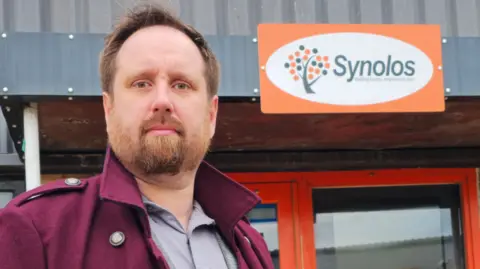 Barry Ingleton, a man with brown hair and a full beard, looks at the camera. He is wearing a maroon coat with an upturned collar over a grey shirt. Behind him is a sign on the front of a building reads "Synolos" next to a stylized symbol representing a tree. 