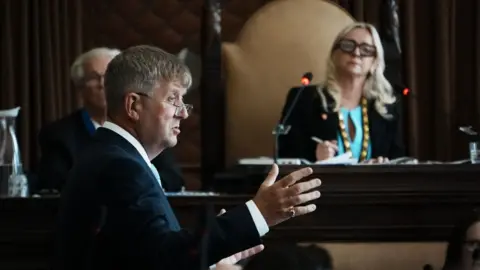 A man in his 50s side-on talking in a council chamber, with his right hand gesticulating as he speaks. Out of focus to the right is the female chairwoman of the debate, wearing a turquoise top.