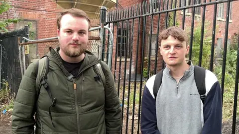 A young man in a green coat wearing a rucksack stands next to another young man wearing a grey, white and blue sweatshirt, who is also wearing a rucksack. The two stand together outside some black railings that are fencing off an abandoned building. 