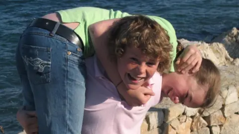 Charlotte Bresnahan Charlotte being carried by Ryan as children. He is wearing a light pink polo top and she is wearing jeans and a green top. The sea is visible in the background over a light stone wall