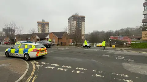 BBC/Tom Oakley Three police officers guard a cordon that stretches from one side of the road to the other. A police car is parked on the left. Three skyscrapers can be seen in the background.