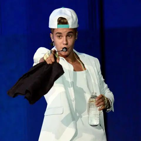 Getty Images Justin Bieber, in an all-white suit with a backwards baseball cap, sings into a headset microphone while holding a towel in his right hand