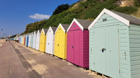 A row of brightly-coloured beach huts stand proudly in the sun in a beach scene. They are all closed.