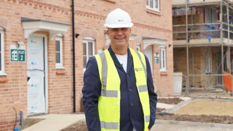 Yorkshire Housing's chief executive Nick Atkin is looking straight into the camera. He is standing on a building site, wearing a bright yellow hi-vis waiscoat, a blue jacket and a white hard-hat.