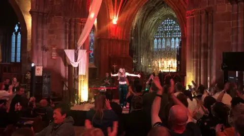 A woman standing on a stage with her arms outstretched. The audience are clapping and smiling