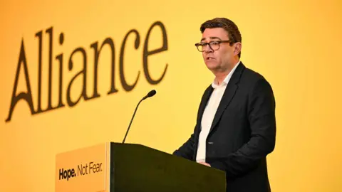 Andy Burnham has short dark hair and stands at a lectern. He is wearing a suit without a tie, and glasses. Behind him is a large yellow screen with 'Alliance' written on it. On the lectern 'Hope. Not Fear.' is written. 