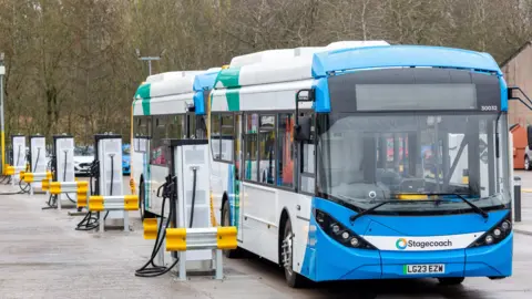 Two electric buses covered in a blue, white, green and yellow livery are parked at depot. They are next to a row of charging points. Trees and bushes are visible in the background.
