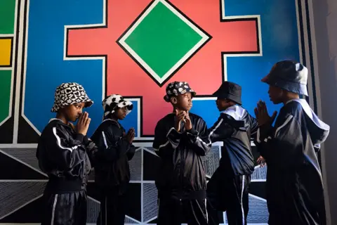ALET PRETORIUS / GALLO IMAGES / GETTY IMAGES Children in dark tracksuits and monochrome bucket hats stand together in room in front of a mural.