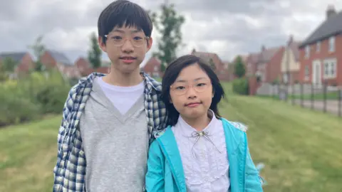 BBC Elias, a boy wearing a grey and white shirt with a blue plaid overshirt, standing next to his sister Ariel, wearing a white shirt and light blue jacket.