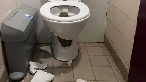 Bethany Lauder A broken toilet bowl in a small tiled public toilet. Large pieces of the broken bowl are on the floor along with smaller shards.