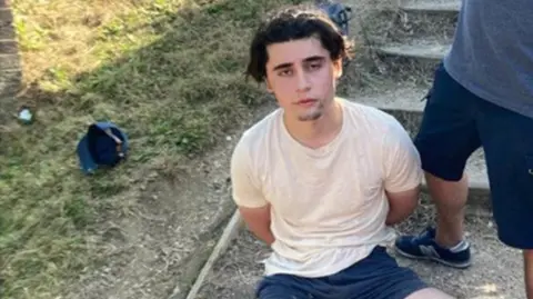 Metropolitan Police Daniel Khalife, wearing a white t-shirt, is pictured sitting on the ground outside with his hands behind his back after his arrest