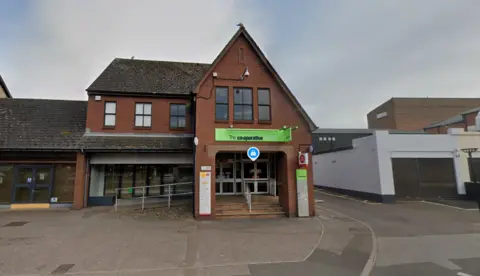 A Google street view image shows the front of the Co-Op on The Avenue. A two-storey red brick building, a green sign reading "The co-operative" sits above the entrance. Beneath it and to the right is a Post Office sign. A lane goes down the right hand side of the shop as we look at it.