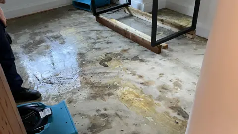 Colin Fletcher A floor is shown to be stained and flooded with grey/brown water. Wooden furniture is seen in the corner. The reflection of puddles can be seen.