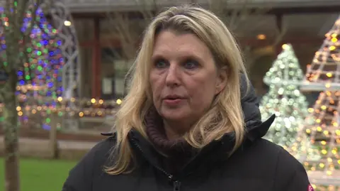 Libby Battaglia stands outside the garden centre, in the background are lit up Christmas trees and trees decorated with fairy lights - they are all slightly out of focus. Libby stands looking slightly off camera as she gives an interview. She has mid-length blonde hair in a side parting and wears a maroon chunky knit turtleneck jumper with a black puffed duvet coat over the top.