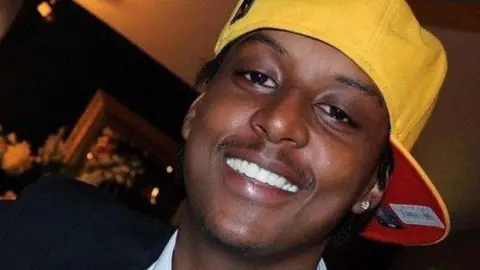 Handout A man wearing a sideways yellow cap and a silver earring in his left ear is smiling at the camera. The image is a close-up so only his head is visible. There appears to be a photo frame in the background but it is blurry.