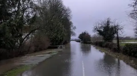 Griffiths Garage A flooded road with trees and countryside on the right-hand side and trees and large white house on the other side. 