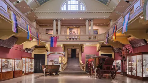 Royal Cornwall Museum Royal Cornwall Museum interior. Stairs in the centre of the museum lead up to a balcony which over looks the inside of the building. It is pink and yellow. Display cupboards line the walls.