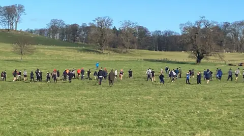 A field with trees on the horizon and a large number of people, in an assortment of clothing.