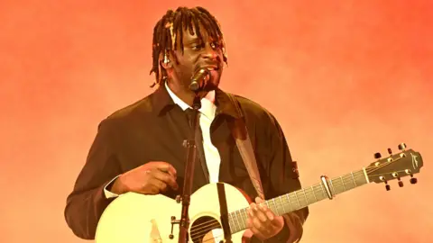 Getty Images Myles Smith is wearing a white shirt and black jacket performing on stage at the 2026 MOBO Awards with a guitar and a microphone. The background behind him is orange. 