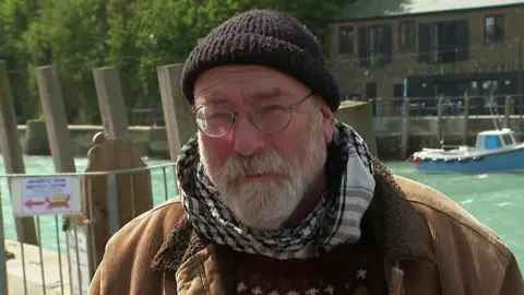 Steve Patterson looking at the camera and stood in front of the water. He is wearing glasses, a hat and a brown jacket with a scarf.