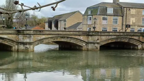 A stock image of Stamford town bridge, with water surrounding the light brown stones of the bridge and light coloured buildings in the background. 