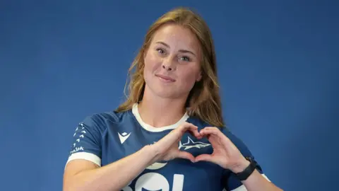 Amelia Hyndman is wearing a blue Sale Sharks kit and creating a heart shape with her hands over the shark logo. She has long, light brown hair and is smiling slightly at the camera. She is standing in front of a blue background for what is a professional shoot.