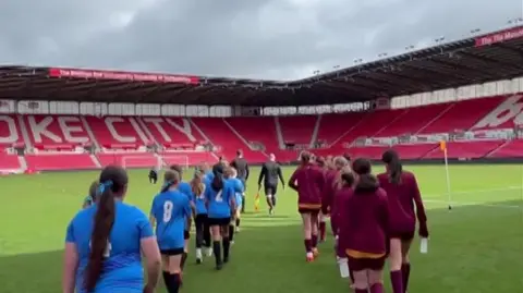 BSG Two teams walking out onto the pitch in single file lines led by a referee. The pitch is surrounded by large red stands.