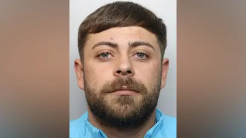 Mugshot of Jordan Woolmer. HE is wearing a light blue jacket and has short brown hair and beard.