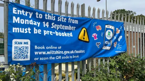 A large blue banner tied to a metal fence. It advertises that the recycling site needs to be booked in future and has a web address and QR code to help people.