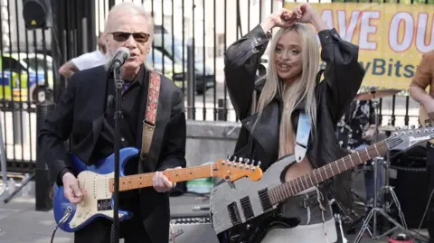 A man with white hair, wearing an all-black outfit and sunglasses, holding a blue electric guitar and singing into a microphone, stands next to a young woman with a breathing tube, long blonde hair, wearing a black leather jacket and a mini skirt and holding a black and white electric guitar. There is a drum set behind them and a yellow banner.