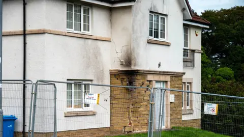 Garry F McHarg The front of a modern white house which has been damaged by a collision causing structural damage to the stone facing. There are cracks in the white render up the front of the two-storey building and scorch marks all the way up. A high metal fence has been erected in front of the property. 