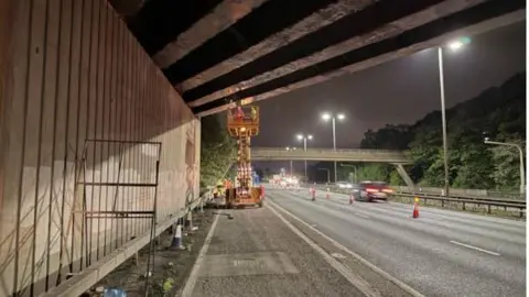 Two workers on a cherry picker in the distance on a hard shoulder under a bridge. A three-lane motorway can be seen next to them with cones marking off two lanes. Blurred cars drive past.
