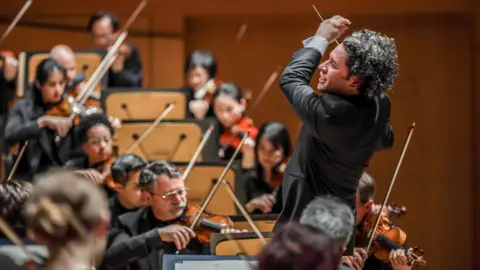 Dustin Downing Dudamel, in formal dress, flamboyantly conducting an orchestra. he has long dark hair.