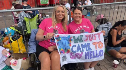 Two women in pink Take That T-shirts in fold out chairs. Their banner says "We just came to say hello".