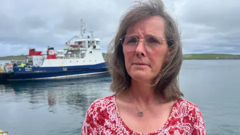 Close-up of a person standing by a harbour wearing round glasses and a red patterned top, with a ferry boat docked on calm water in the background under a cloudy sky.