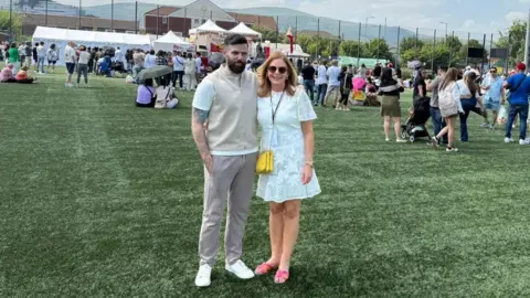 A man and woman stand on a green Astroturf pitch, about a hundred people stand behind them. A large stage can be seen in the background. The man wears a white t-shirt with a cream vest over it with grey trousers and white shoes. He has shirt brown hair and a beard. The woman wears a small white dress, yellow handbag and Pink sandals. She wears brown sunglasses and has long brown hair.