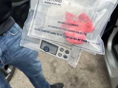 Photo of two police evidence bags being held by someone in blue jeans. One bag contains a red wrapper with white powder; the other has a set of digital scales.