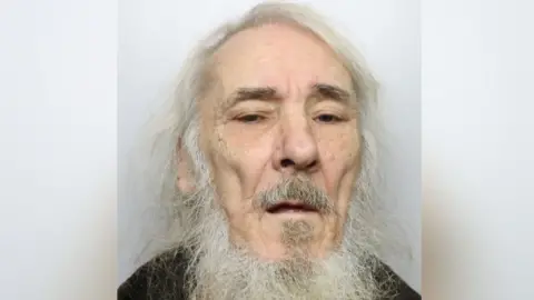 An elderly man with long white hair and beard. He is standing against a grey background wearing a black top.