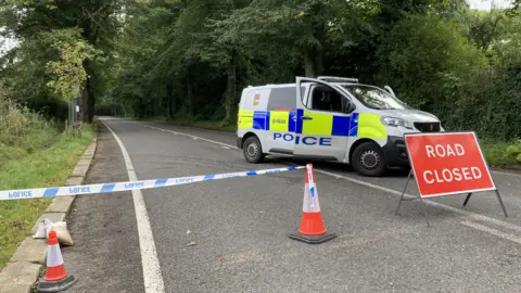 A country road which goes through Rivelin Valley in Sheffield. The photo is taken from just behind some police tape. A sign in line with the tape reads "road closed". Just behind the sign is a police van with the front doors open. There is no debris or signs of an incident on the road other than this.