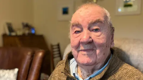 Richard Aldred is looking into the camera and smiling. He is wearing a brown striped jacket.