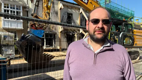 A man in a purple jumper stands in front of a grey fence and a mechanical digger.