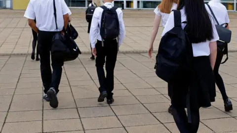 A generic images of the backs of schoolchildren in dark trousers, shoes, skirts and tights and white shirts, walking with rucksacks on stone paving towards a building.
