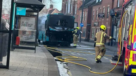 Sarah Gayton A fire engine is stopped in the middle of a street, with firefighters carrying hoses towards a bus. The bus is blackened at the back and there is smoke coming from the side of it. In the distance is a row of building as well as road signage.