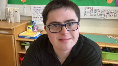 Ben has black hair, black glasses and a black shirt on. He sits in a classroom in front of a noticeboard.