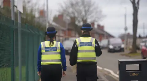 Two people in hi-viz vests with the words Police Community Support Officer on the back are walking away from the camera, on a residential street