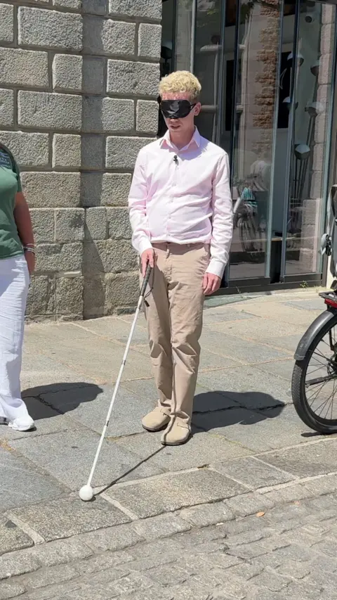 Man in a pink shirt and cream trousers with a black blindfold and holding a white cane