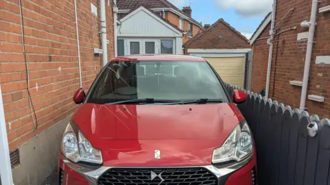 Handout A red Citroen car sitting in a driveway with a grey gate on the left and brick wall on the right.