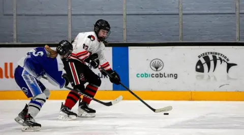 Alan Blakeley Molly is seen playing ice hockey avoiding a player as she controls the puck. She is wearing a white jersey and black trousers with red and black striped socks, her opponent is in blue and white.