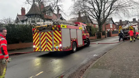 A fireman stands by a fire engine with its lights on in the street near the convent with firemen moving towards another engine